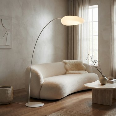 A modern floor lamp with a curved silver metal arm and a glowing oval shade stands beside a cream boucle sofa in a softly lit living room with wood floors and sheer curtains.