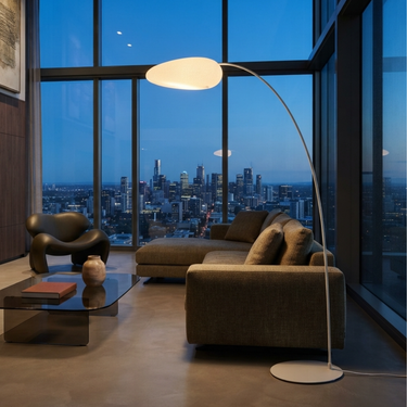A modern living room with a large white arched floor lamp beside a brown sectional sofa, with a city skyline visible through tall windows at dusk.